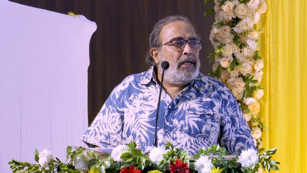 ফুলে সাজানো মঞ্চে মাইক্রোফোনের সামনে চশমা ও নীল ছাপা জামা পরা দাড়িওয়ালা এক ব্যক্তি।