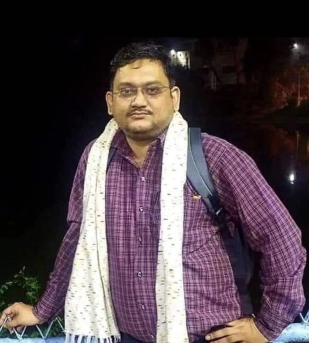চশমা, বেগুনি চেক জামা ও সাদা উত্তরীয় পরা এক ব্যক্তির প্রতিকৃতি; কাঁধে ব্যাগ।